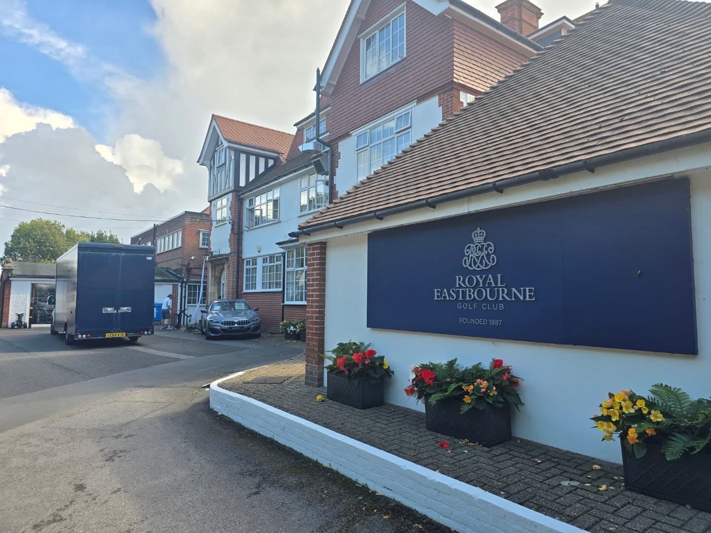 Entrance to Royal Eastbourne Golf Club with a blue sign and potted flowers. A large brick building, car, and truck are in the background under a cloudy sky.
