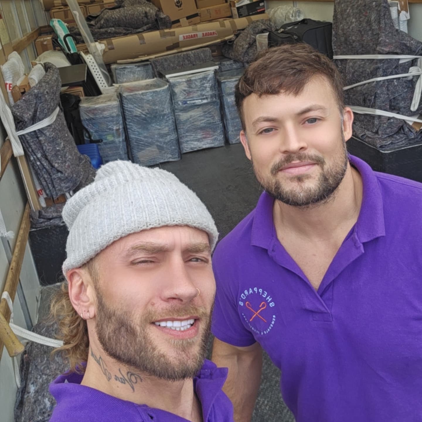 Two smiling men in purple shirts take a selfie inside a moving van filled with wrapped furniture and boxes, conveying a cheerful and friendly atmosphere.
